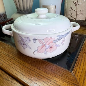May include: A white ceramic casserole dish with a lid and handles. The dish is decorated with a floral pattern in shades of pink, purple, and gray. The dish is sitting on a wooden surface.