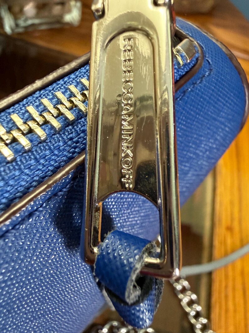 K&ouml;nnte beinhalten: Eine leuchtend blaue, strukturierte Handtasche mit einem polierten, silberfarbenen Metall-Rei&szlig;verschlussanh&auml;nger, der mit dem Text REBECCAMINKOFF graviert ist. Die Tasche hat eine passende blaue Lederlasche und eine silberne Metallkette.