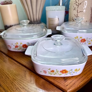 May include: A set of four white square casserole dishes with clear glass lids. Each dish features a floral design with orange, yellow, and blue flowers. The dishes are on a wooden table.