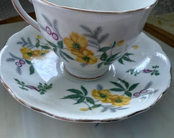 May include: White teacup and saucer set with a floral design. The teacup and saucer feature yellow flowers, green leaves, and purple berries. The rims are trimmed with gold. The teacup has a curved handle.