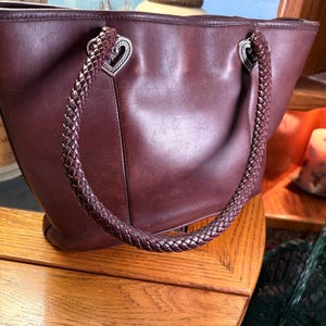 May include: A dark brown leather tote bag featuring two braided leather handles attached with silver-toned metal hardware shaped like hearts. The bag has a smooth, matte finish and a structured silhouette with visible stitching along the sides.