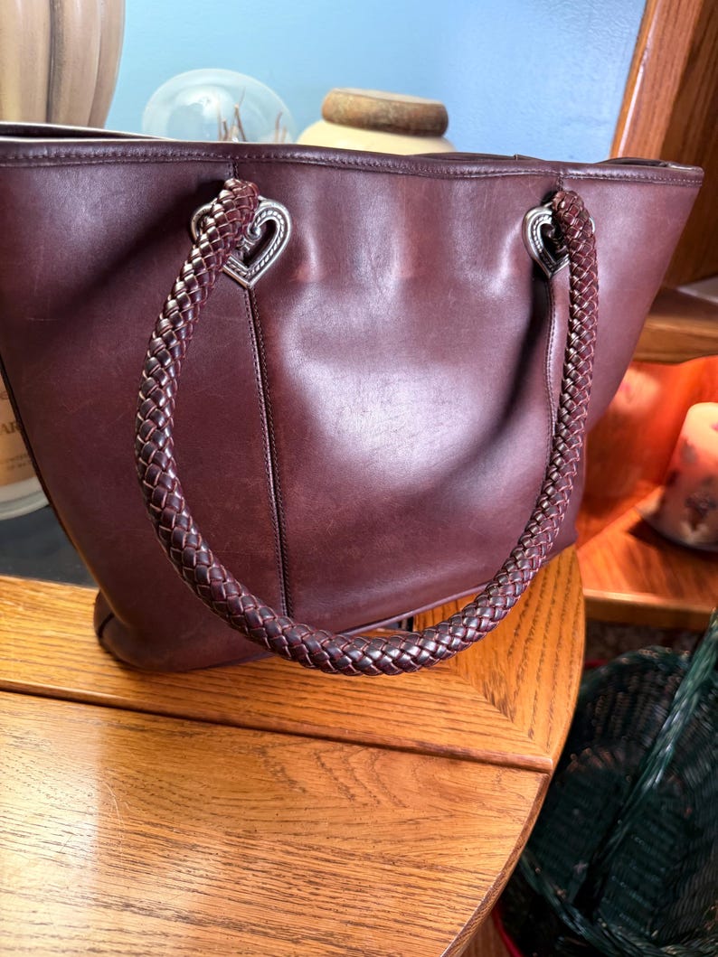 May include: A dark brown leather tote bag featuring two braided leather handles attached with silver-toned metal hardware shaped like hearts. The bag has a smooth, matte finish and a structured silhouette with visible stitching along the sides.
