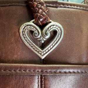 May include: A dark brown leather handbag featuring a prominent silver-toned metal heart-shaped charm with intricate embossed scrollwork details. The bag includes a thick, braided dark brown leather handle attached directly to the heart ornament.
