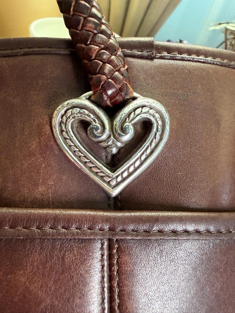 May include: A dark brown leather handbag featuring a prominent silver-toned metal heart-shaped charm with intricate embossed scrollwork details. The bag includes a thick, braided dark brown leather handle attached directly to the heart ornament.