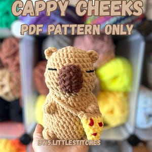 May include: A crocheted amigurumi toy of a brown capybara holding a yellow and red crocheted pizza slice. The toy is sitting in a hand. The background is a shelf with colorful yarn balls.