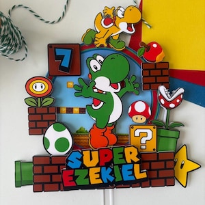 May include: A colorful cake topper featuring Yoshi from Super Mario, with the text "SUPER EZEKIEL". The design includes a green Yoshi, a yellow Yoshi, a red apple, a mushroom, and a star. The background has a blue sky and brick details.
