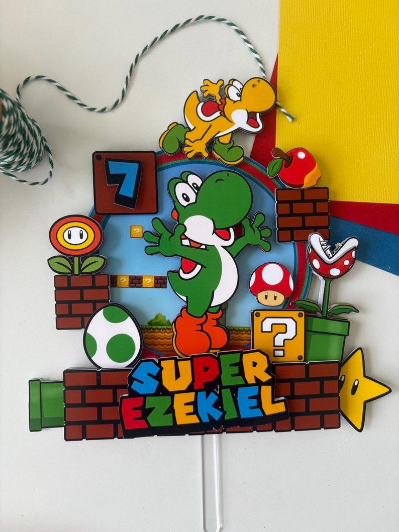 May include: A colorful cake topper featuring Yoshi from Super Mario, with the text "SUPER EZEKIEL". The design includes a green Yoshi, a yellow Yoshi, a red apple, a mushroom, and a star. The background has a blue sky and brick details.