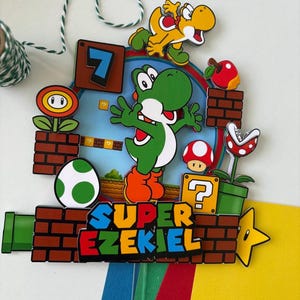 May include: A colorful Super Mario-themed decoration featuring Yoshi, a yellow dinosaur, and other game elements. The design includes a green pipe, brick blocks, a question mark box, and the text "SUPER EZEKIEL".