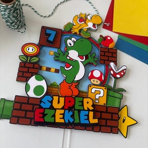 May include: A colorful cake topper featuring a Super Mario Bros. theme. The design includes Yoshi, a yellow dinosaur, a red mushroom, and the text "SUPER EZEKIEL". The topper is made of layered paper and has a white stick for insertion.