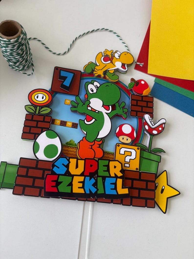 May include: A colorful cake topper featuring a Super Mario Bros. theme. The design includes Yoshi, a yellow dinosaur, a red mushroom, and the text "SUPER EZEKIEL". The topper is made of layered paper and has a white stick for insertion.