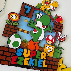 May include: A colorful Super Mario-themed cake topper featuring Yoshi, a yellow dinosaur, a red apple, a mushroom, and a Piranha Plant. The topper includes the text "SUPER EZEKIEL" and the number 7. The design is set against a brick background.