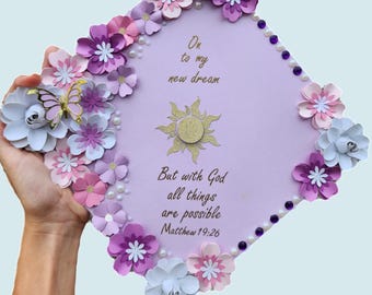 May include: Purple graduation cap decorated with paper flowers in shades of pink, white, and purple. The cap has a quote in gold lettering: "On to my new dream. But with God all things are possible. Matthew 19:26"