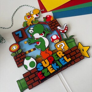 May include: A colorful cake topper featuring Yoshi from Super Mario. The design includes a green Yoshi, a yellow Yoshi, a question mark block, a star, and the text "SUPER EZEKIEL". The topper is attached to a clear plastic stick.