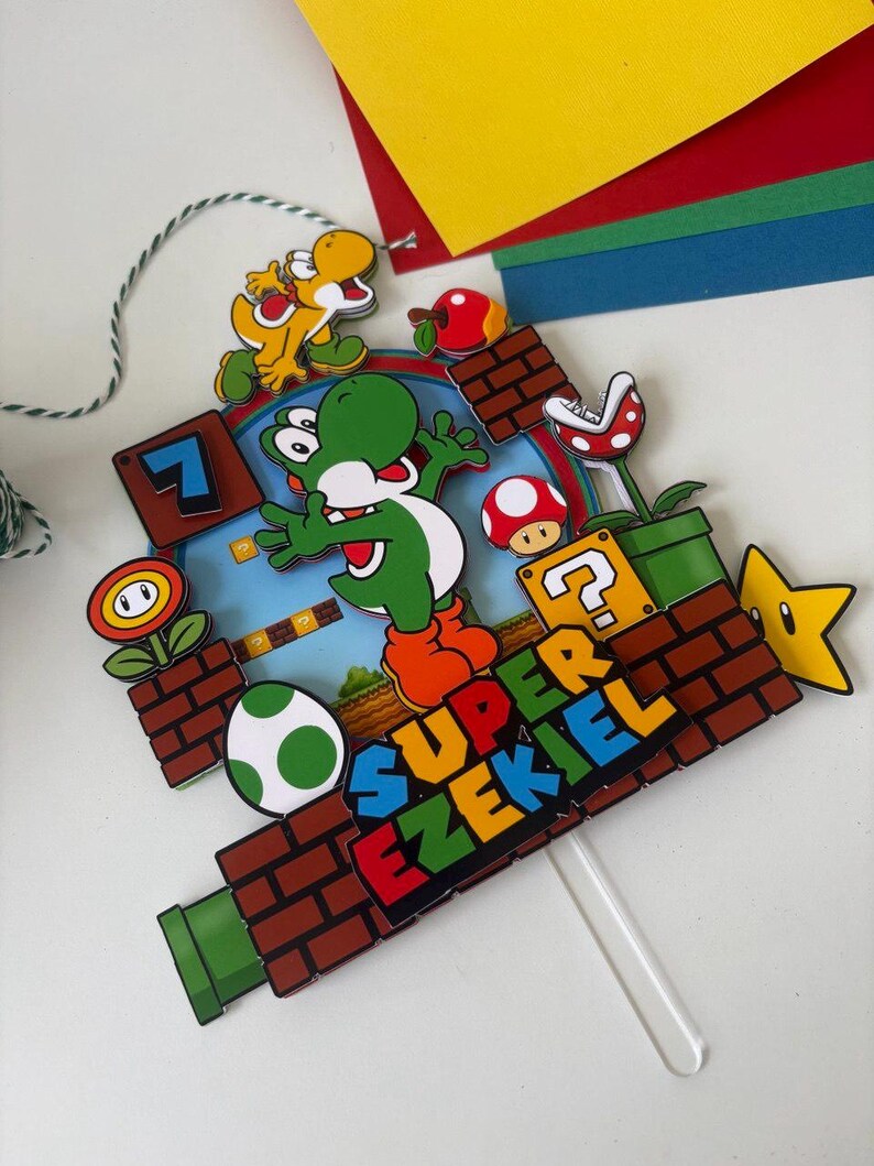 May include: A colorful cake topper featuring Yoshi from Super Mario. The design includes a green Yoshi, a yellow Yoshi, a question mark block, a star, and the text "SUPER EZEKIEL". The topper is attached to a clear plastic stick.