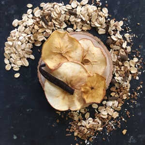 May include: Dried apple slices arranged on a wooden surface with a vanilla bean. The apple slices are a light brown color and have a natural, rustic look. The background is a dark gray surface with scattered oats.