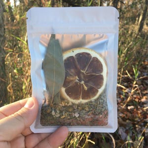 May include: A clear, resealable bag containing a dried lemon slice, a bay leaf, and a mix of herbs and spices. The contents are visible through the transparent packaging. The background is blurred, with natural light and foliage.
