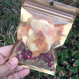 May include: A clear plastic bag with a gold zipper closure contains dried fruit and flower petals. The bag is filled with dried pear slices, apple slices, orange peel, and red flower petals.