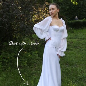 May include: A white dress with a corset-style bodice and a long skirt with a train. The dress has long, sheer sleeves with ruffled cuffs. The model is standing in a grassy area with trees in the background. Text on the image reads 'skirt with a train'.