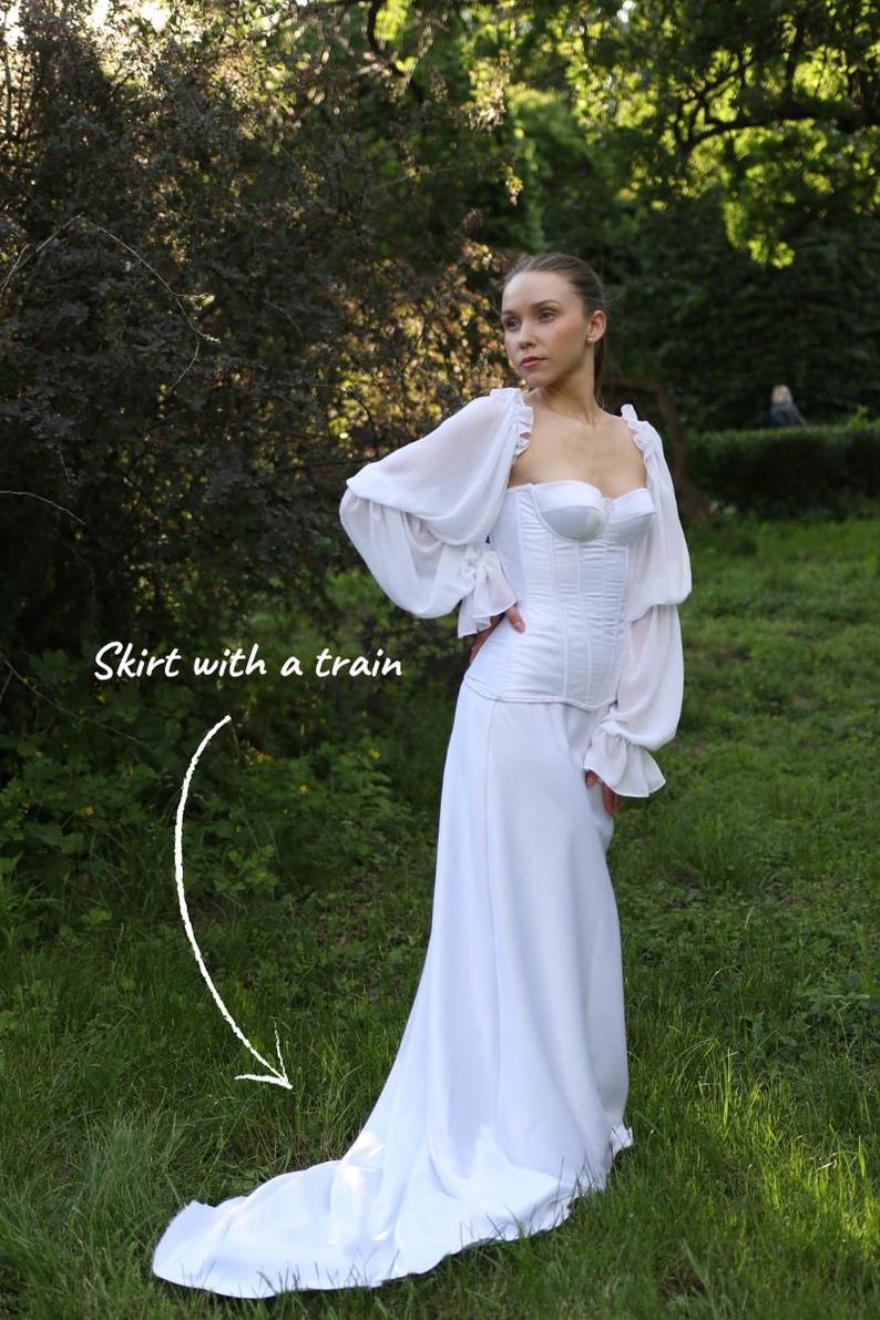 May include: A white dress with a corset-style bodice and a long skirt with a train. The dress has long, sheer sleeves with ruffled cuffs. The model is standing in a grassy area with trees in the background. Text on the image reads 'skirt with a train'.