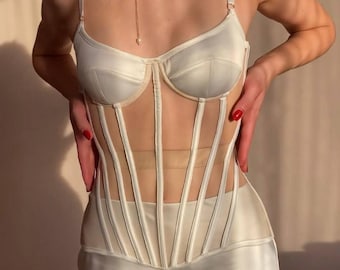Può includere: Top a corsetto color avorio con pannello trasparente e dettagli di stecche verticali. Il top presenta uno scollo a cuore e spalline regolabili. È visibile anche una collana di perle con catena dorata e un pendente a perla singola.