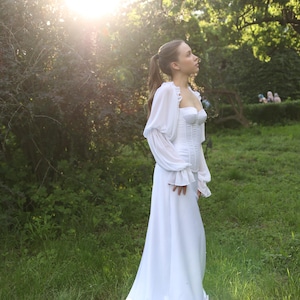 May include: A white wedding dress with a long train. The dress has a fitted bodice with a sweetheart neckline and long, sheer sleeves. The dress is worn outdoors in a grassy area with sunlight in the background. The dress is flowing and elegant, perfect for a romantic wedding.