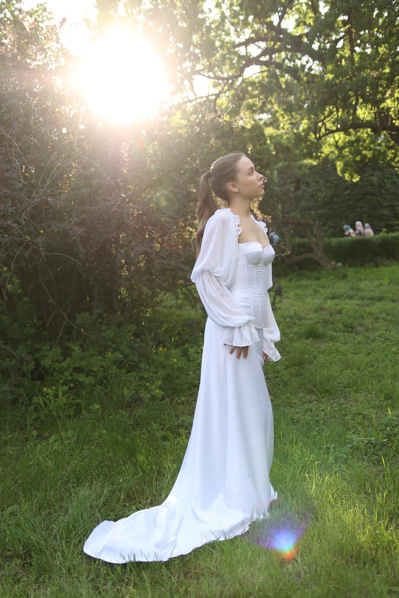 May include: A white wedding dress with a long train. The dress has a fitted bodice with a sweetheart neckline and long, sheer sleeves. The dress is worn outdoors in a grassy area with sunlight in the background. The dress is flowing and elegant, perfect for a romantic wedding.