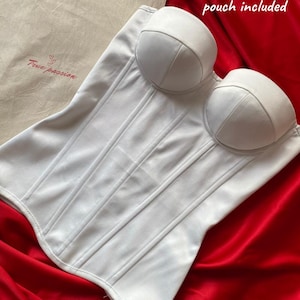 May include: White corset with vertical boning details and padded cups. The corset is displayed on a red satin fabric. A branded beige storage pouch with the text 'True passion' is included. The image also includes the text 'Branded storage pouch included'.