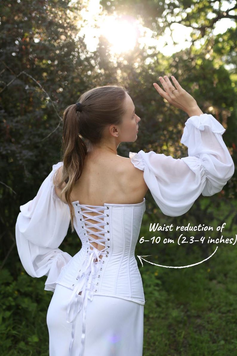 May include: A white corset with a lace-up back, paired with a matching white skirt and long-sleeved, off-the-shoulder top. The corset is cinched, and the image includes text indicating a waist reduction of 6-10 cm (2-3-4 inches). The outfit is worn outdoors.