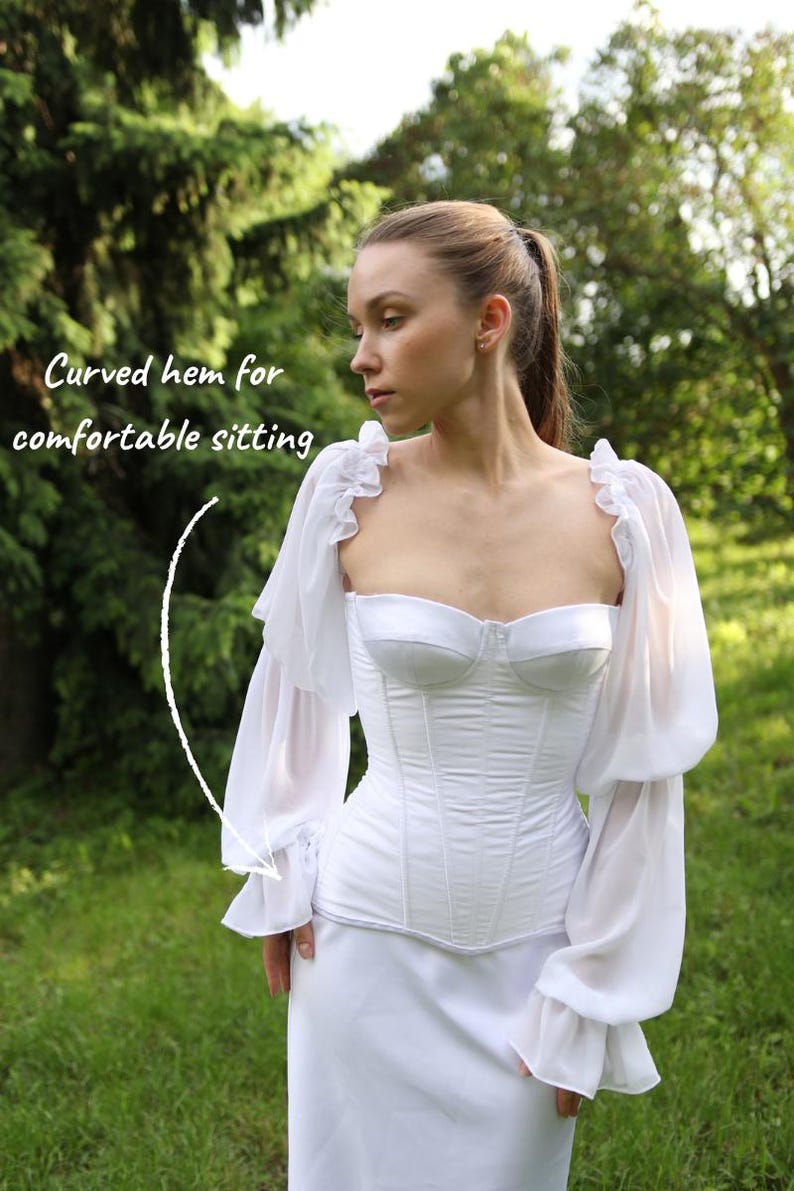 May include: A white corset-style top with a sweetheart neckline and boning details. The top has long, sheer sleeves with ruffled shoulders and cuffs. The model is wearing a matching white skirt. The text on the image says 'Curved hem for comfortable sitting'.