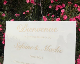 May include: White sign with gold lettering announcing a wedding. The sign reads "Bienvenue au mariage religieux de Safoine & Maelis 09.09.2023 Et nous vous avons créés par paires » S78 : V8"