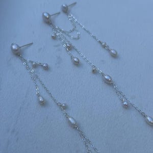 May include: Elegant silver earrings with dangling chains and lustrous white pearls. The earrings feature a stud at the top with multiple chains of varying lengths, each adorned with small and large pearls. A delicate and sophisticated jewelry piece.