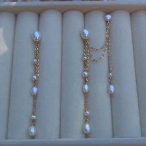 May include: A pair of gold chain earrings featuring multiple small white freshwater pearls. The left earring consists of a single vertical chain with pearls, while the right earring features a double-chain design with pearls and a connected stud.