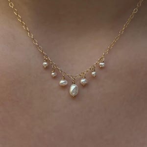 Könnte beinhalten: Eine zarte goldene Halskette mit kleinen, herabhängenden Perlen. Die Halskette hat eine feine Kette und mehrere kleine Perlen unterschiedlicher Größe, die daran hängen. Die Perlen sind weiß und haben ein glänzendes Finish.