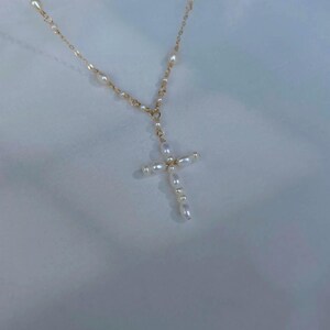 May include: A gold chain necklace featuring small, white, oval-shaped freshwater pearls spaced along the links, culminating in a cross-shaped pendant constructed entirely from similar white pearls arranged in a vertical and horizontal pattern.