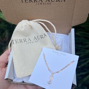 K&ouml;nnte beinhalten: Eine kleine wei&szlig;e Schachtel mit einer goldenen Halskette darin. Die Halskette hat eine zarte Kette und einen kleinen Perlenanh&auml;nger. Die Schachtel ist mit "Terra Aura Jewelry" beschriftet.