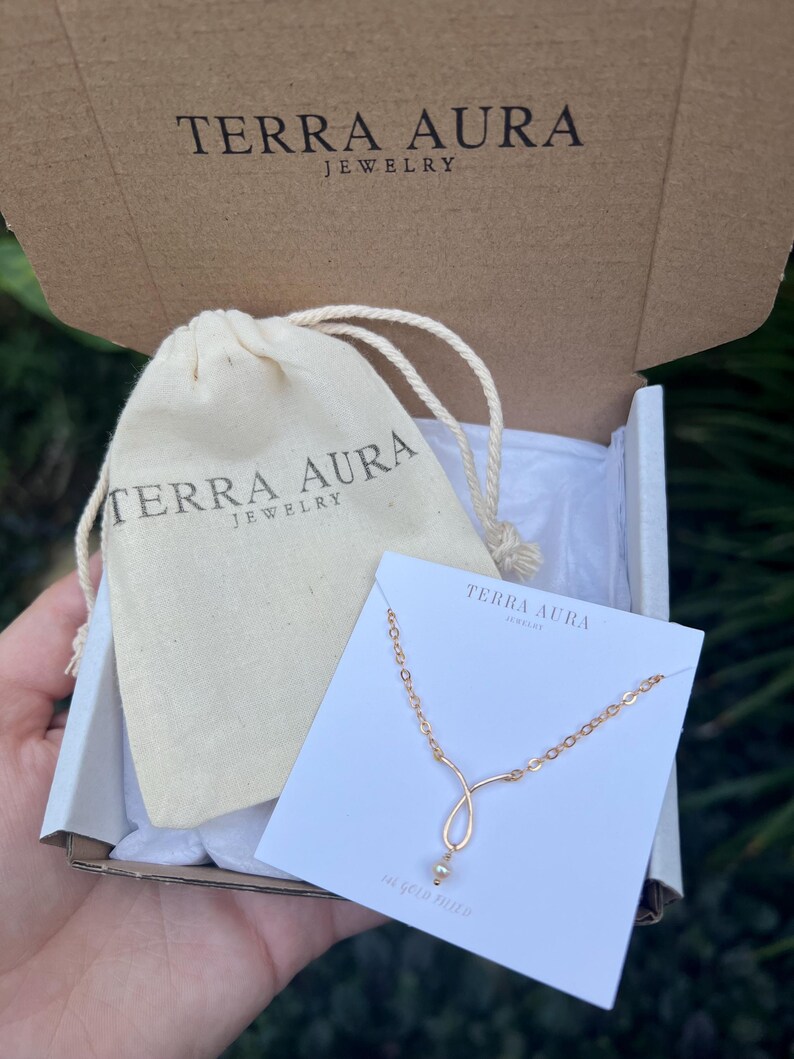K&ouml;nnte beinhalten: Eine kleine wei&szlig;e Schachtel mit einer goldenen Halskette darin. Die Halskette hat eine zarte Kette und einen kleinen Perlenanh&auml;nger. Die Schachtel ist mit "Terra Aura Jewelry" beschriftet.