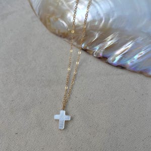May include: A delicate gold chain necklace with a small white cross pendant. The necklace is displayed on a white fabric background.