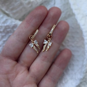May include: A pair of gold-colored hoop earrings with a bow-shaped charm. The hoops and charms are encrusted with small, clear gemstones. The earrings are displayed against a soft, textured background.