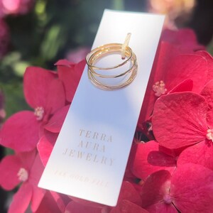K&ouml;nnte beinhalten: Ein Satz von vier goldfarbenen Stapelringen auf einer wei&szlig;en Karte mit dem Text "Terra Aura Jewelry 14k Gold Fill". Die Karte liegt auf einem Strau&szlig; rosa Bl&uuml;ten.