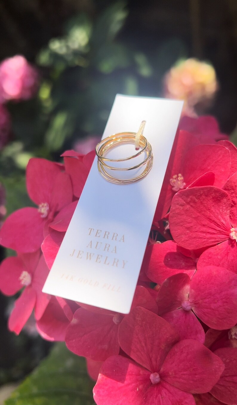 K&ouml;nnte beinhalten: Ein Satz von vier goldfarbenen Stapelringen auf einer wei&szlig;en Karte mit dem Text "Terra Aura Jewelry 14k Gold Fill". Die Karte liegt auf einem Strau&szlig; rosa Bl&uuml;ten.