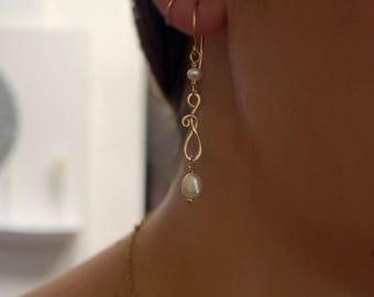 May include: Gold-toned drop earrings with a delicate, swirling design. Each earring features a small, round pearl and a larger, teardrop-shaped pearl at the bottom. The earrings are shown against a blurred background.