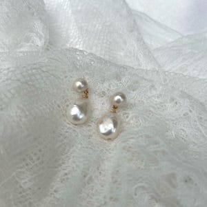 May include: A pair of drop earrings featuring two white pearls of varying sizes connected by a thin gold-toned metal wire. The smaller round pearl sits above the larger, slightly irregular teardrop-shaped pearl, creating a dangling design.