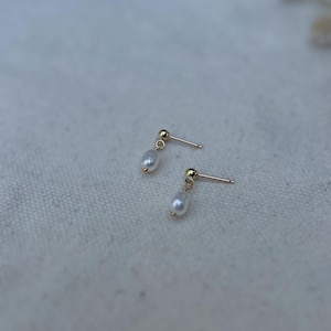 May include: A pair of gold stud earrings, each featuring a small, pear-shaped pearl pendant. The earrings are set against a light, textured background. These earrings are a delicate piece of jewellery.