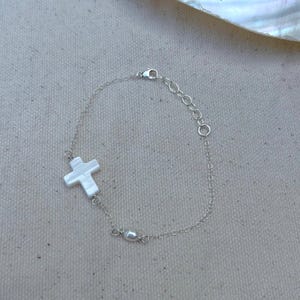 May include: A silver chain bracelet with a white mother of pearl cross charm and a single white pearl charm.