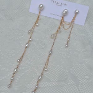 May include: Elegant gold-toned earrings featuring a pearl stud and cascading pearl accents. The earrings are attached to a delicate chain. The earrings are displayed on a white surface with a Terra Aura Jewelry tag.