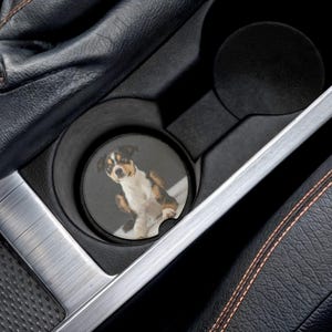 May include: A car cup holder coaster featuring a dog. The coaster has a black background with a full-color image of a dog with brown, black, and white fur. The coaster is placed in a car's cup holder, surrounded by black and silver trim.
