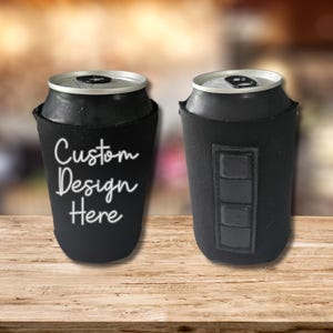 May include: Two black can coolers, each holding a silver can. One cooler features the text "Custom Design Here" in white script. The other cooler has a black rectangular patch with three stitched squares.