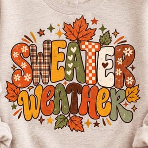 May include: A light heather gray sweatshirt featuring the text Sweater Weather in large, colorful, retro-style letters. The design includes orange, green, and brown autumn leaves, small white flowers, checkered patterns, and tiny star shapes.