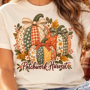 May include: A cream-coloured t-shirt featuring a graphic of three pumpkins with patchwork patterns in orange, green, and yellow. Each pumpkin is topped with a bow. The text "Patchwork Harvest" is printed in a brown script font below the pumpkins.