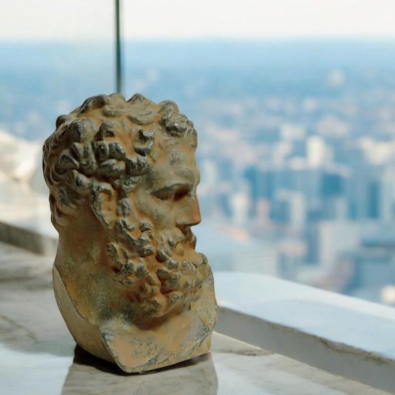 May include: A weathered, gray stone bust of a man with a beard and curly hair. The bust is facing to the right and is set against a blurry background of a city skyline.